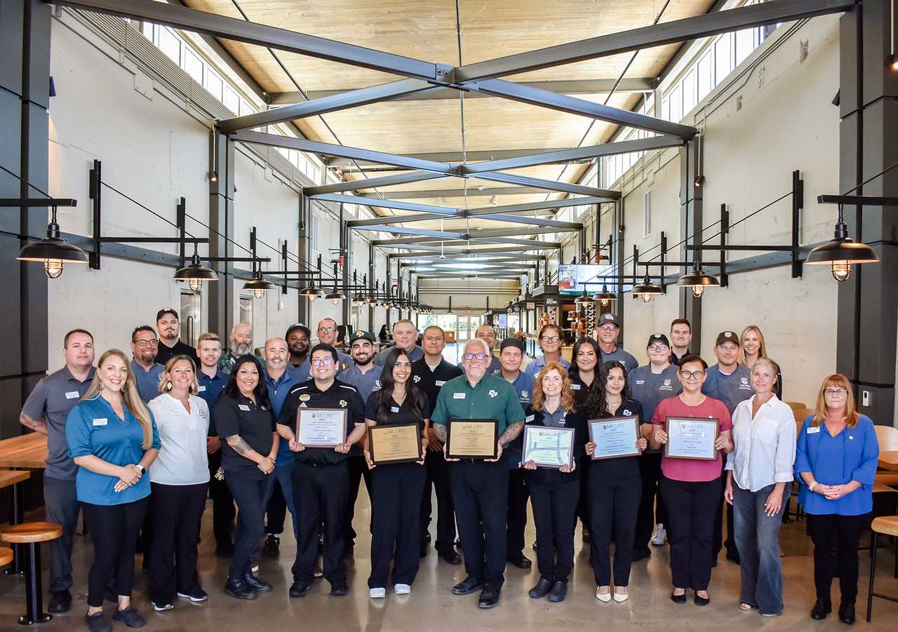 Group of Cal Poly staff members standing together inside a modern dining hall, holding awards and certificates while smiling for a photo.