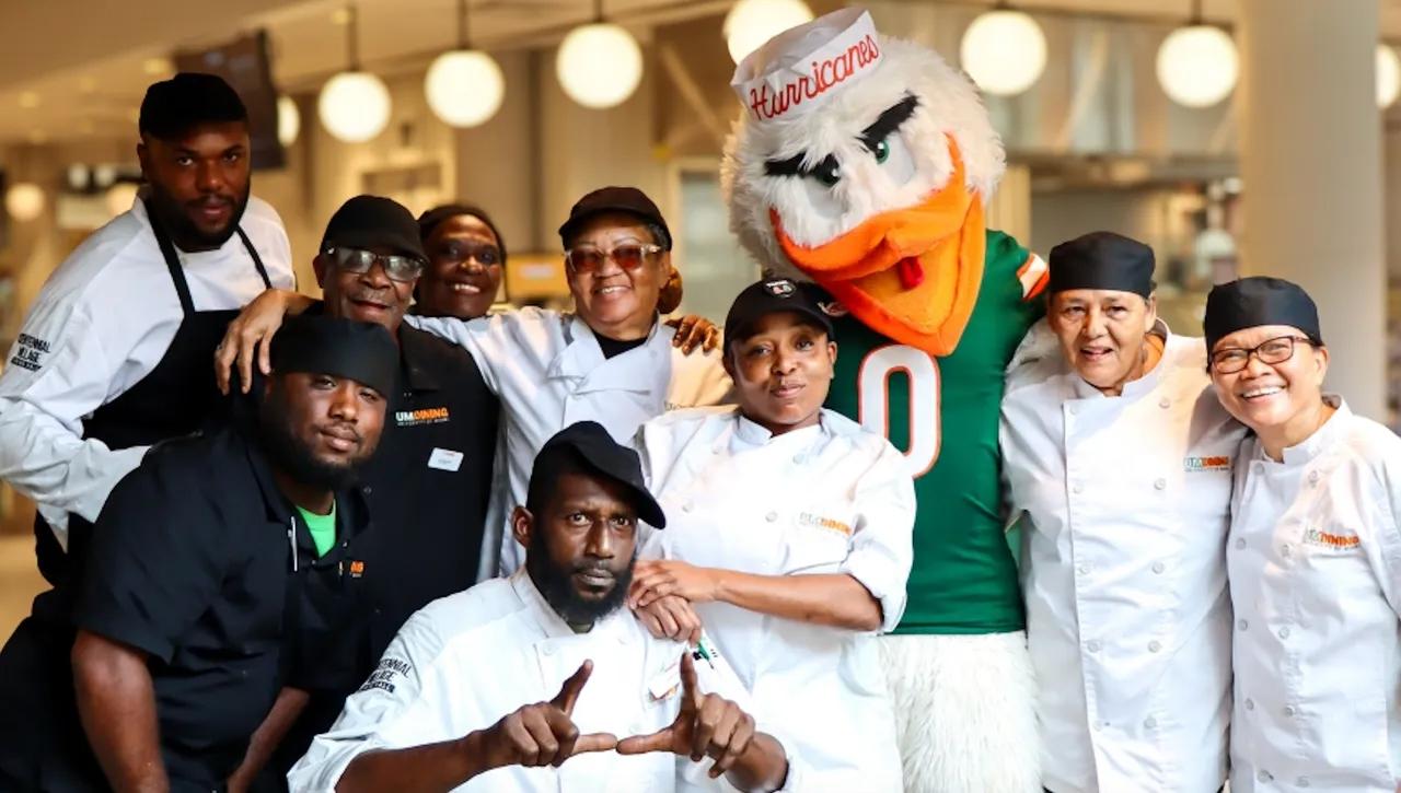 A group of smiling kitchen staff members pose together with a university mascot dressed in green and orange, wearing a “Hurricanes” hat, inside a brightly lit dining facility.