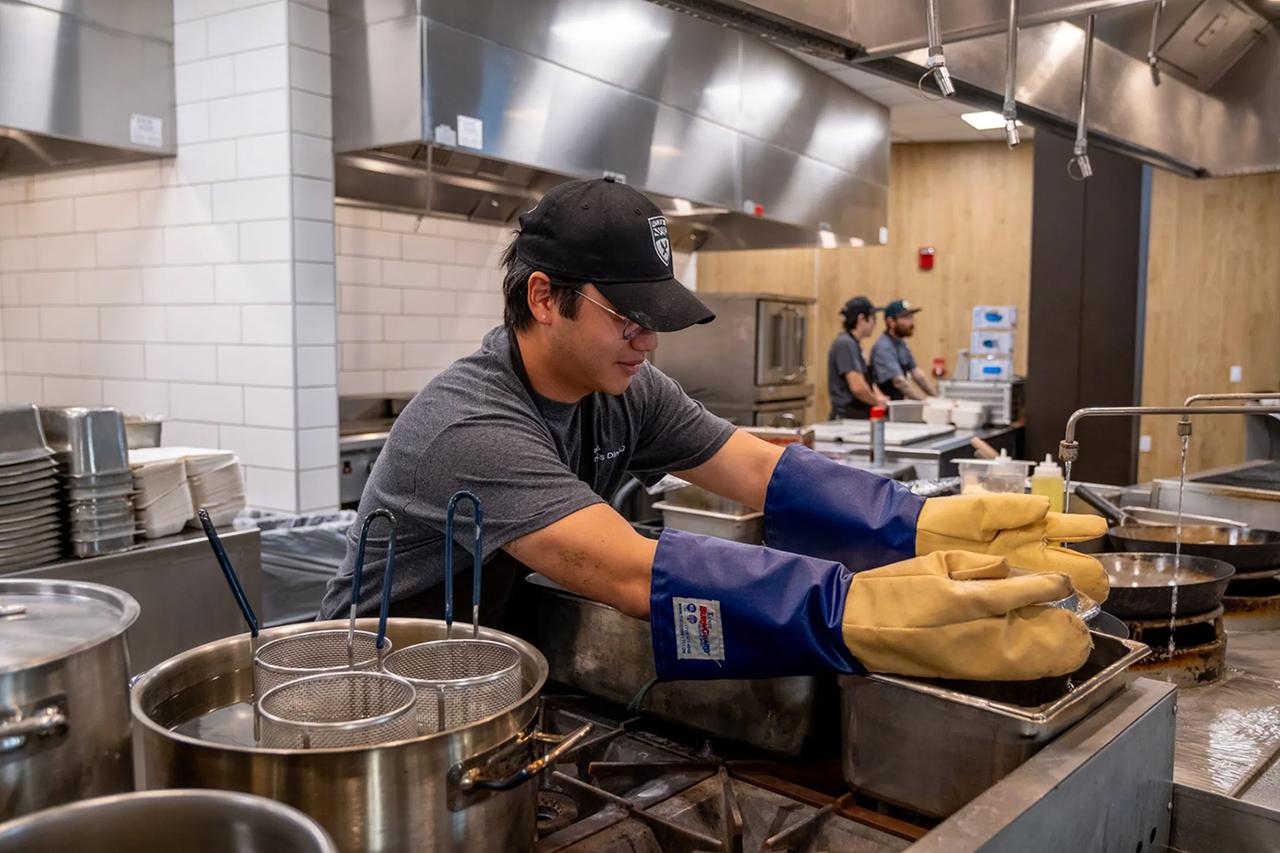 A Cal Poly Dining employee wearing protective gloves and a hat with the Cal Poly logo works in a commercial kitchen, preparing food near large stainless steel pots and fry baskets, with coworkers visible in the background.