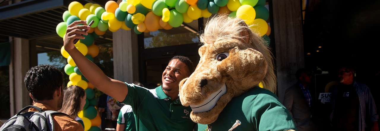 A student takes a selfie with the Cal Poly Mustang mascot in front of a building decorated with green and yellow balloons during a campus celebration.