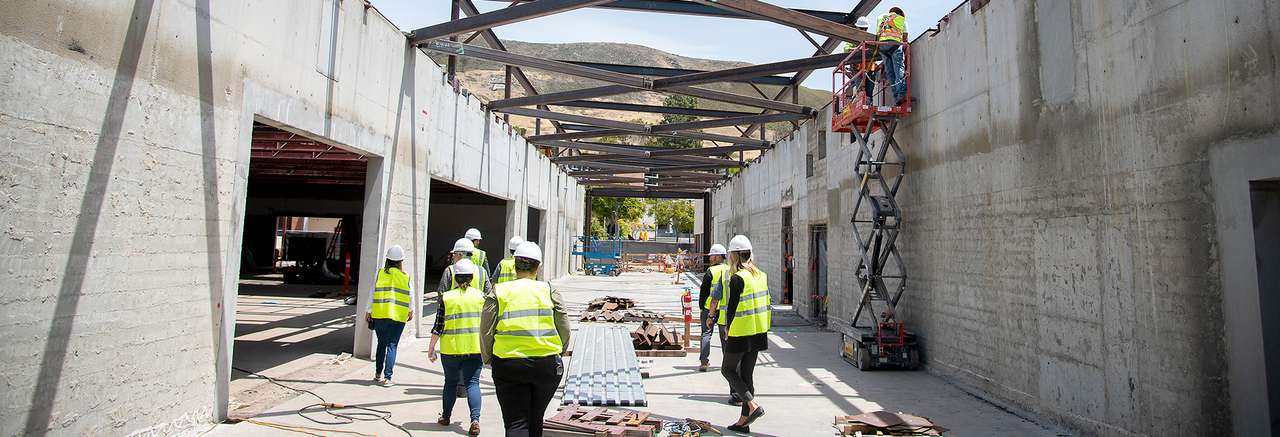 Construction workers and project team members wearing hard hats and safety vests inspect a large concrete building under construction, with exposed steel beams and equipment on site.