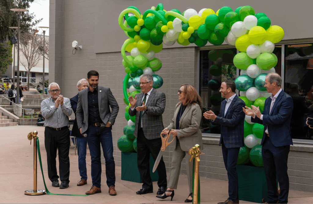 A group of people stand under a green and yellow balloon arch during a ribbon-cutting ceremony; one woman holds oversized scissors while others applaud.