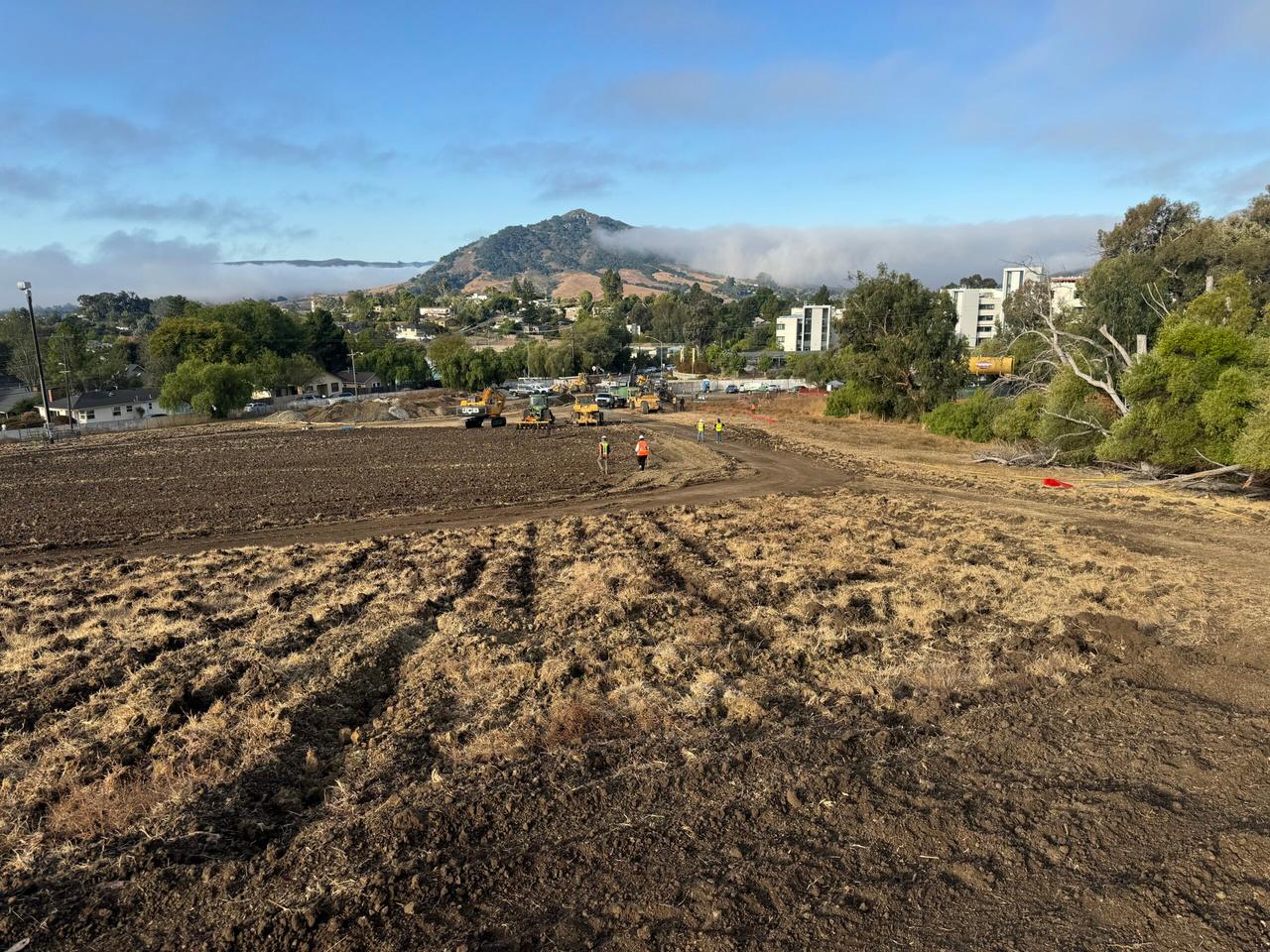 Construction crews working on a large dirt field at Cal Poly San Luis Obispo with equipment and vehicles in the background, surrounded by trees, campus buildings, and hills under a partly cloudy sky.